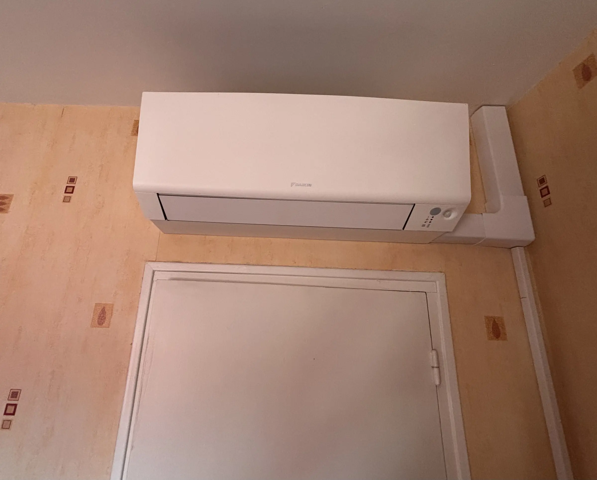 Climatiseur mural blanc posé au-dessus d’une porte blanche, une goulotte blanche descend dans l’angle d’un mur au papier peint orangé
