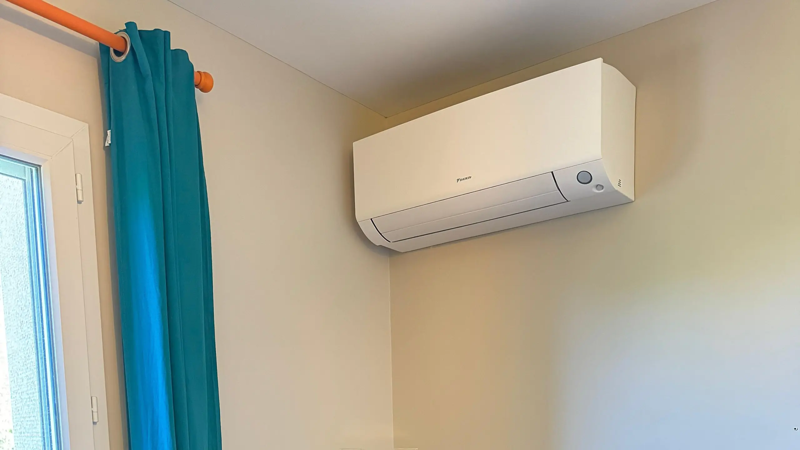 Climatiseur mural blanc fixé en haut d’un mur crème, à côté d’une fenêtre au rideau bleu canard, une vitrine à figurines en dessous