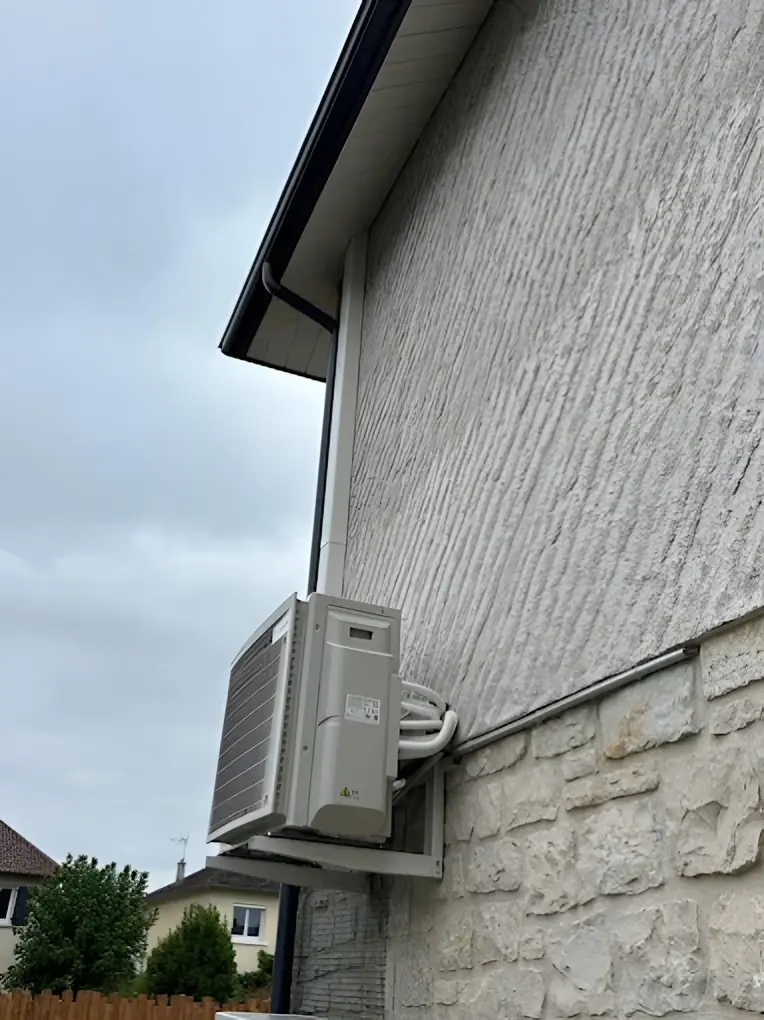Groupe extérieur de climatisation vu de profil sur une console fixée à une façade crépie, soubassement en pierre, maisons voisines au fond