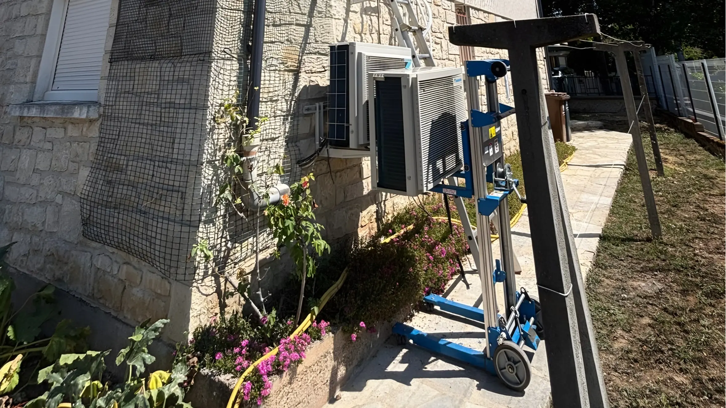 Façade crépie à soubassement de pierre : un groupe extérieur de climatisation sur un lève-charge bleu, à côté d’un autre déjà fixé sur ses équerres, une échelle contre le mur