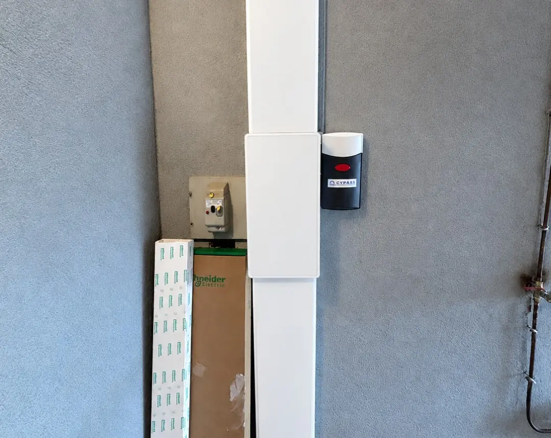 Gaine technique blanche verticale fixée sur un mur gris, boîtier noir à côté, ancien disjoncteur et cartons de matériel à gauche