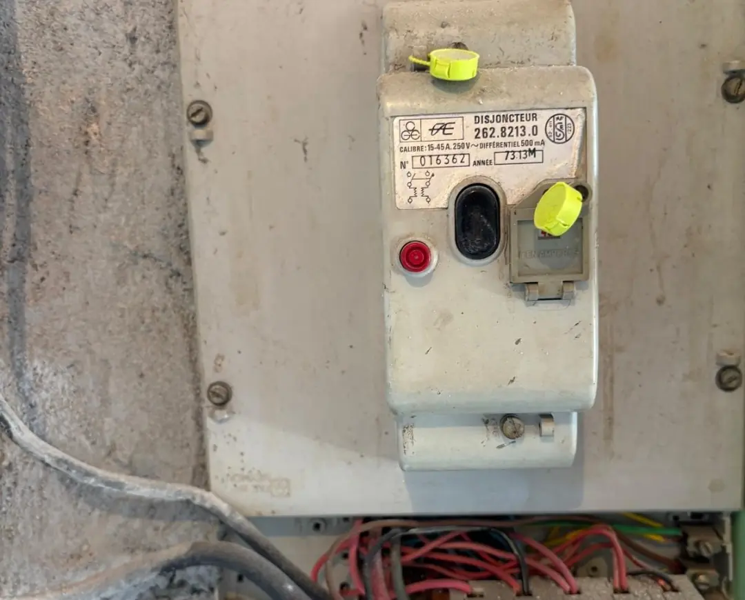 Ancien disjoncteur de branchement beige au bouton noir, étiquette « année 73 », fils rouges et noirs en vrac en dessous